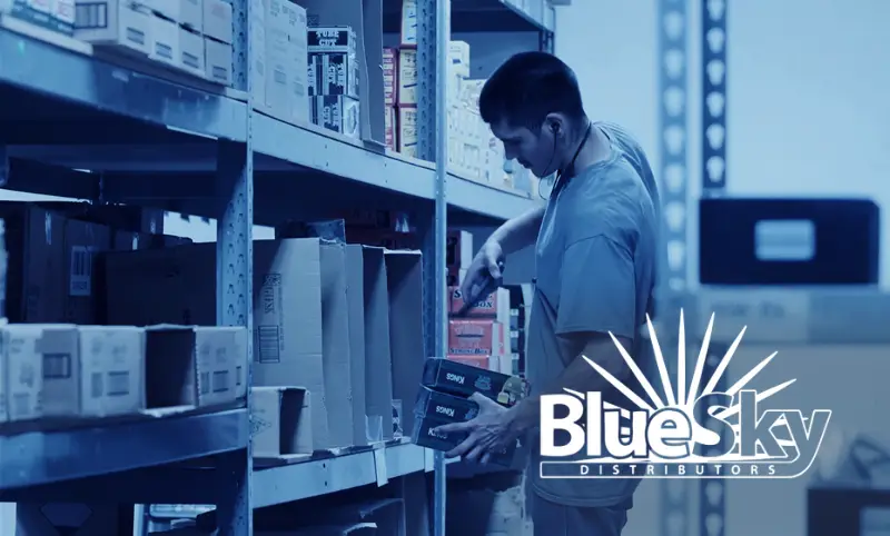 Blue Sky Distribution logo with background of a warehouse worker using a mobile device with warehouse management system for NetSuite to scan an item on a shelf
