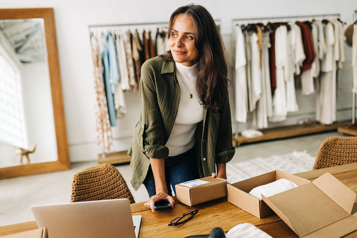 A small business owner thinks about adding multiple ecommerce marketplaces while standing at a packing station with parcels and a laptop computer in a room with racks of clothing and a mirror.