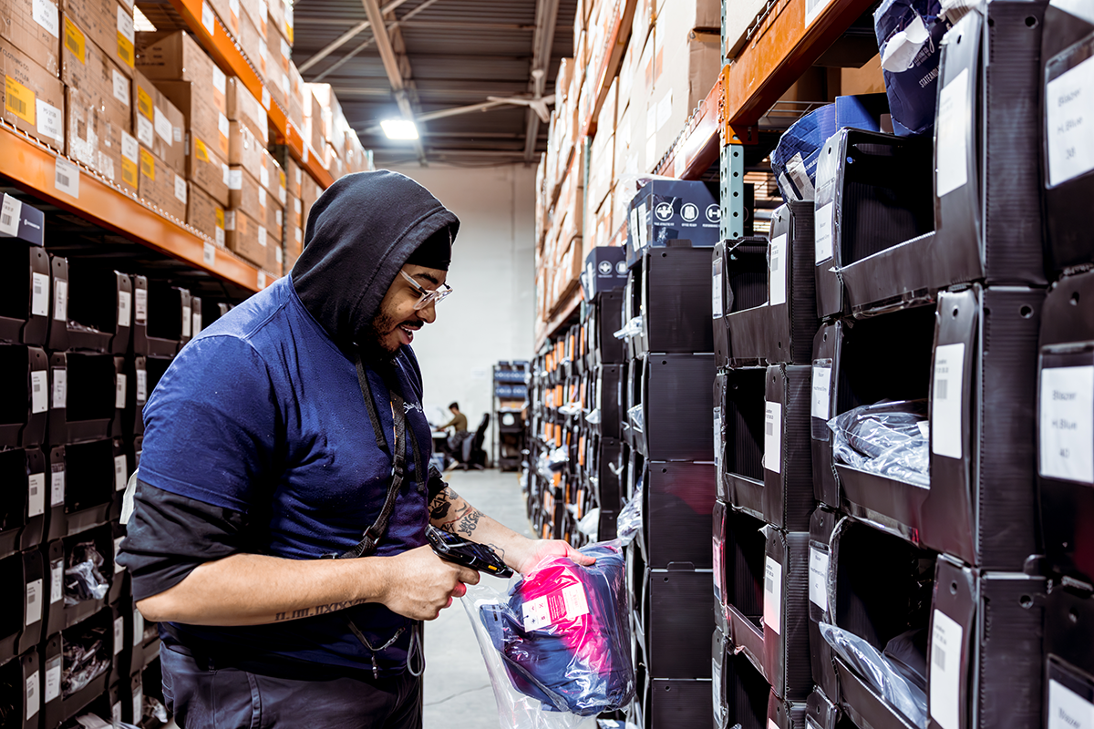 A worker in a warehouse aisle holds an item and scans it using a mobile barcode scanner to ensure multichannel inventory sync as orders are picked.